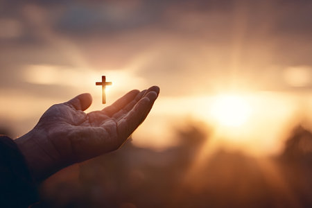 Human hand holding a glowing Christian cross against a warm sunset sky, symbolizing faith, hope, prayer, devotion and spiritual guidance in golden hour lightの写真素材