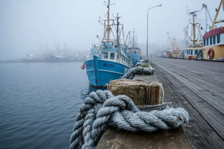 Fishing boats moored to a weathered wooden pier with thick ropes in a foggy harbor, evoking quiet maritime industry, solitude, and cold coastal atmosphereの写真素材