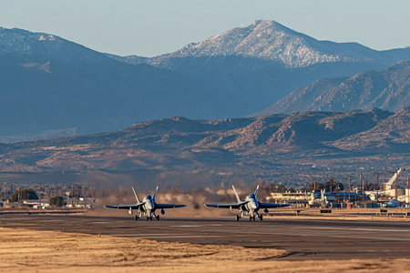 Two fighter jets are accelerating on an asphalt runway, kicking up dust and exhaust, with a desert...