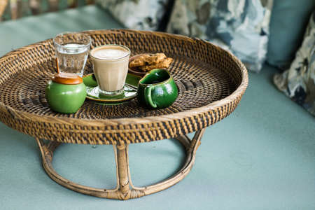Rattan tray with cappuccino, milk gravy boat, sugar bowl, glass of water and cookies on blue sofa with pillows. Lazy morning concept.の写真素材