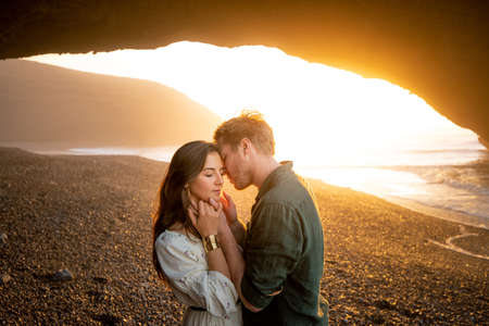 Romantic couple standing near natural huge arch on the beach. Handsome man and attractive woman enjoying sunset in Morocco.の写真素材