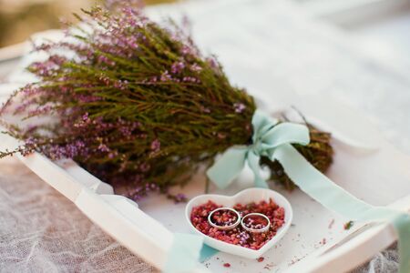Platinum or silver wedding rings on the heart shaped white plate with pink heather flowers and wedding bouquetの写真素材