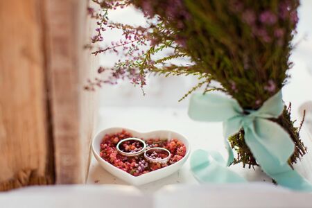 Platinum or silver wedding rings on the heart shaped white plate with pink heather flowers, vintage...