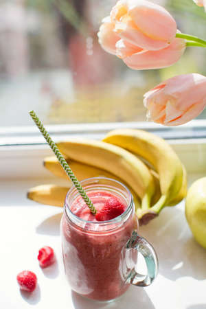 Healthy diet smoothie with strawberry, banana and apples in glass mason jar. Smoothie and bouquet of tulips on windowsillの写真素材