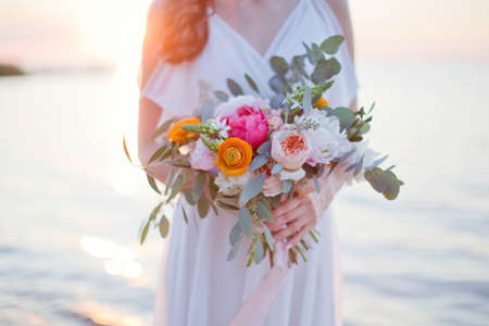 Bride holds wedding bouquet with beautiful flowers and pink silk ribbons. Young woman with flowers standing near seaの写真素材