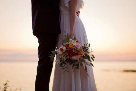 Young man and woman with bright bouquet stand near the sea at sunset lightsの写真素材