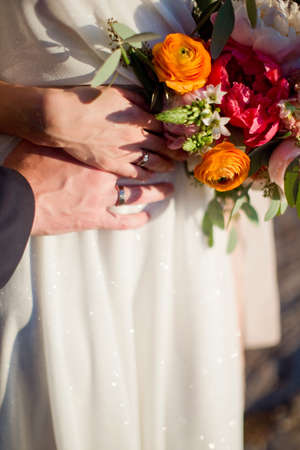 Beautiful bright bouquet with peonies, ranunculus and roses and woman's and man's hands with wedding ringsの写真素材