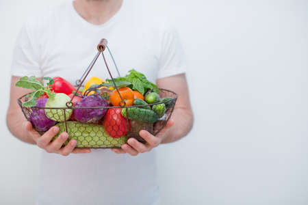 Caucasian man holds metalic vasket with fresh vegetables on a white background. Farm organic vegetables delivery. Food deliver with basket with peppers, tomatoes, cucumbers, basil leaves and kohlrabiの写真素材