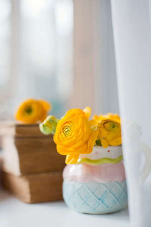 Orange ranunculus in cute small pitcher and stack of vintage books on a windowsill