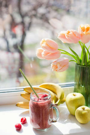 Healthy diet smoothie with strawberry, banana and apples in glass mason jar. Smoothie and bouquet of tulips on windowsillの写真素材