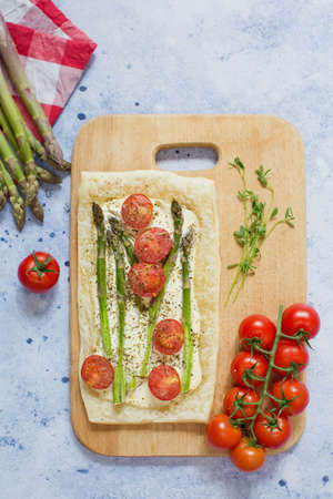 Pie with green asparagus, cream cheese and tomatoes on a wooden cutting board on a light blue backgroundの写真素材
