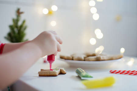 Cute toddler boy decorates Christmas cookies with colorful icing on a white table near Christmas tree with lights. Christmas cooking conceptの写真素材