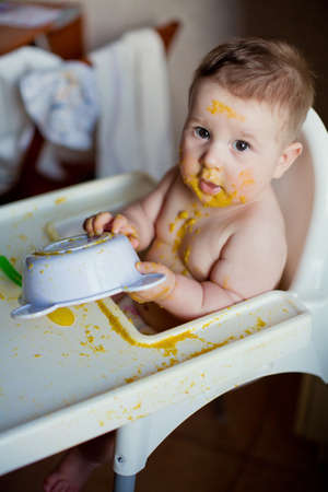 Cute boy with dirty messy face sitting in high chair eating orange vegetable puree with fingers