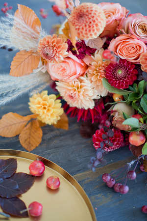 Beautiful autumn bouquet with orange and red flowers and berries. Autumn bouquet and golden tray with small apples on a wooden tableの写真素材