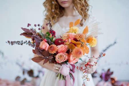 Bride holds beautiful autumn bouquet with orange and red flowers and berries. Autumn bouquet with ribbons in bride's handsの写真素材
