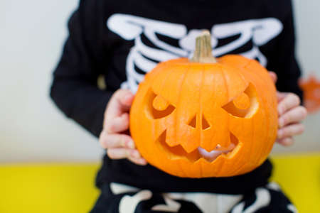 Toddler caucasian boy in skeleton costume holds curved pumpkin and sits on the table with Halloween food and decorationsの写真素材
