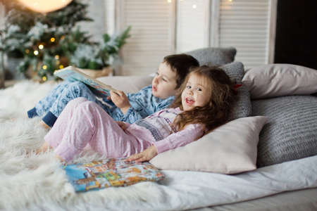 Toddler boy and girl in pyjamas lying on the bed and read books near Christmas tree in light Christmas interior. Cute christmas kidsの写真素材