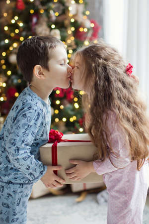 Toddler boy and girl in pyjamas kissing and hold gift boxes near Christmas tree in light Christmas interior. Cute christmas kidsの写真素材