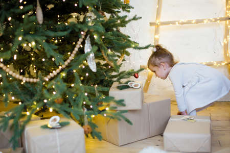Cute little girl in a white dress stands near Christmas tree with decorations and lightsの写真素材
