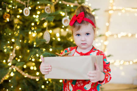 Cute little girl in a red pajamas holds gift box and stands near Christmas tree with decorations and lightsの写真素材