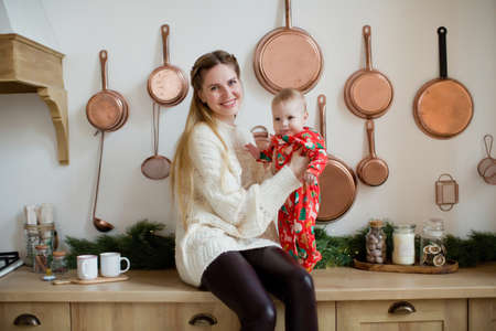 Young mother and little baby boy in red pajamas sitting on the table at Christmas kitchen with Christmas decorations and lightsの写真素材