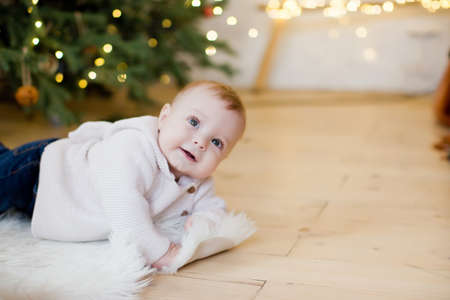 Cute little baby boy is lying near Christmas tree with decorations and lights. Cozy light interior with Christmas decorations and lightsの写真素材