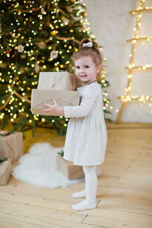 Cute little girl in a white dress with gift boxes stands near Christmas tree with decorations and...