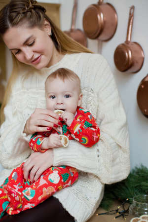 Young mother and little baby boy in red pajamas sitting on the table at Christmas kitchen with Christmas decorations and lightsの写真素材