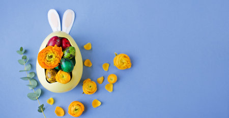 Easter eggs, orange flowers and paper bunny ears in the yellow bowl on a blue background. Easter colorful conceptの写真素材