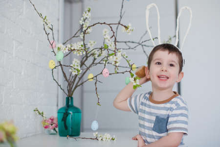 Caucasian toddler boy with funny bunny ears stands near easter tree with colorful eggs on a white...
