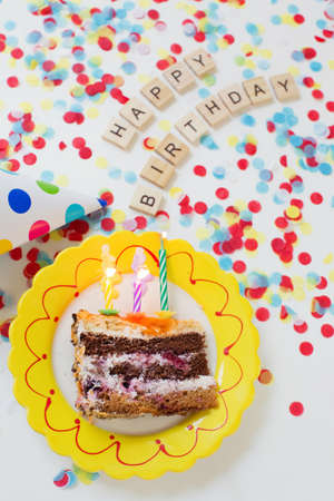 Birthday background with tasty cake on a plate, paper party hat, colorful confetti and wooden sign...