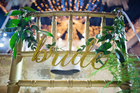 Golden chairs with sign bride and groom with green ruscus twigs. Event hall with golden decorations and tropic palm leavesの写真素材