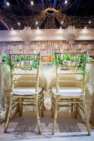Golden chairs with sign bride and groom with green ruscus twigs. Event hall with golden decorations and tropic palm leavesの写真素材