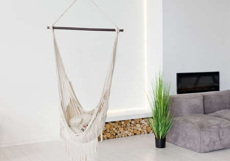 White hanging swing chair in light modern interior with white walls, wooden logs and houseplant