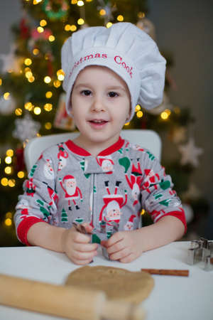 Cute toddler boy making Christmas cookies on a white table near Christmas tree with lights. Christmas cooking conceptの写真素材