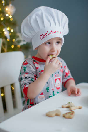 Cute toddler boy eats Christmas cookies on a white table near Christmas tree with lights. Christmas cooking conceptの写真素材