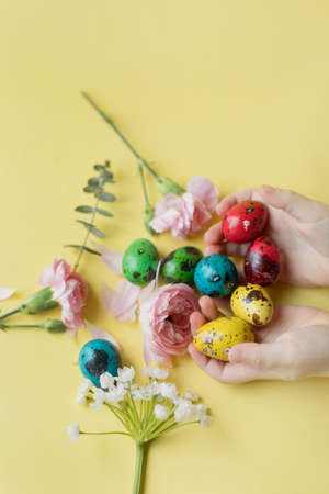 Easter eggs and flowers on a yellow background. Childs hand touch small quail colorful eggs. Easter background with pastel colors details. spring holiday conceptの写真素材