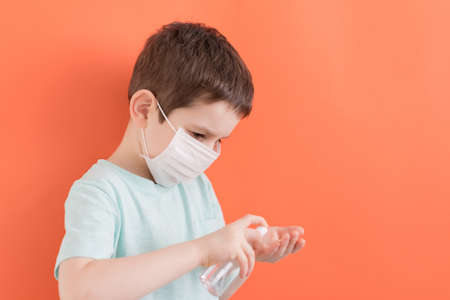Preschool caucasian boy holds white medical mask and sanitizer on orange background.の写真素材