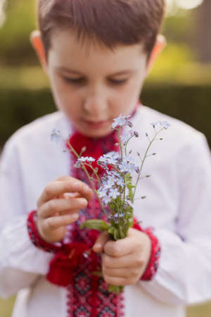 Caucasian boy in traditional ukrainian cloth vyshyvanka at the blossom garden holds dandelion. Ukraine ethnic costume, kid wear patriotic clothの写真素材