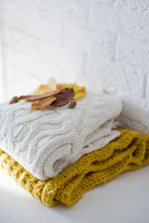 Knit sweaters pile stack and autumn leaves near white brick wall. Cozy fall autumn concept