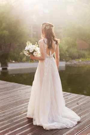 Bride with white wedding bouquet. Woman in white dress standing on a wooden floor outdoors at sunset light and holding bouquet with peonies, lisianthus eustoma and ruscus. Happy wedding conceptの写真素材