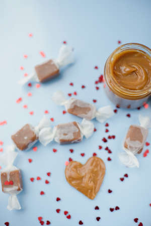Jar with caramel sauce, caramel candies in paper wrapping, heart shaped caramel decoration on a blue...