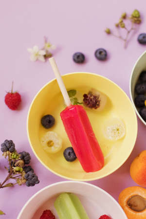 Bright colorful ice lolly in colorful bowls with berries and ice cubes on a light violet background. Tasty ice pops close upの写真素材