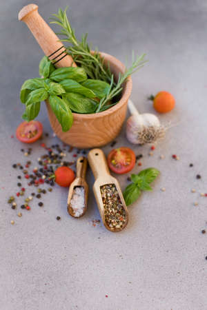 Wooden mortar and pestle with fresh rosemary and basil, spices and tomatoes on a grey concrete background with copy space. Spices and fresh kitchen herbs conceptの写真素材