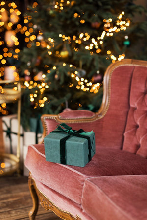 Green gift box with ribbon on a vintage pink sofa near Christmas tree with decorations and lightsの写真素材