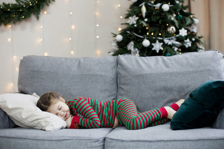 Preschool caucasian girl in Christmas pyjamas slepping on a grey sofa near Christmas tree with decorationsの写真素材