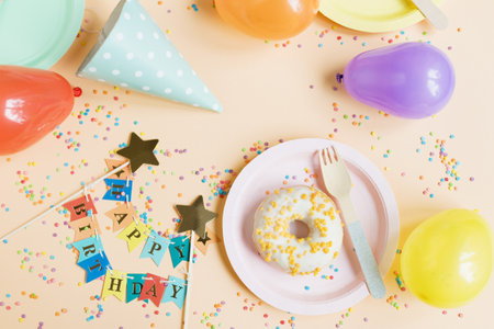 Tasty donut on a pink plate near colorful balloons, birthday hat, happy birthday garland on a beige background with confetti. Happy birthday party table setting concept with copy spaceの写真素材