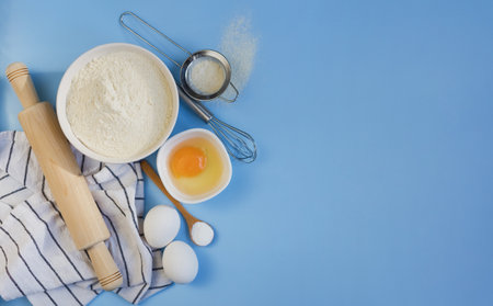 Ingredients for baking on blue background. Flour in bowl, eggs, baking powder and kitchen utensils. Ingredients banner with copy spaceの写真素材