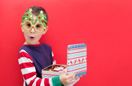 Caucasian boy in funny glasses holding vintage metal box with vintage decorations on a red...