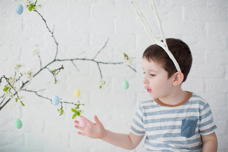 Caucasian toddler boy with funny bunny ears near easter tree with colorful eggs on a white...